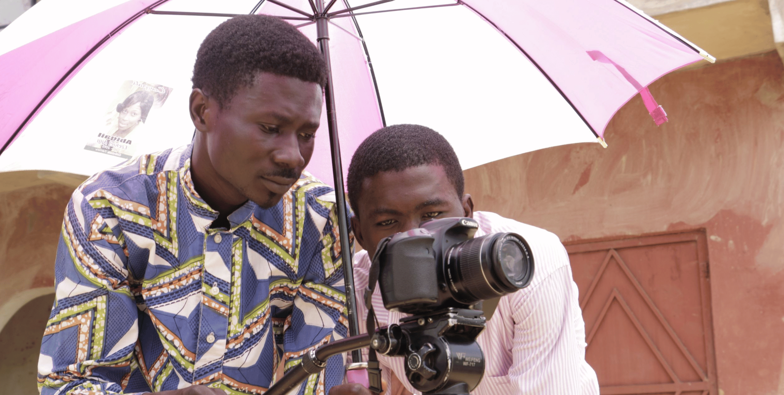 Two men hold a video camera under an umbrella. They are filming something out of frame.