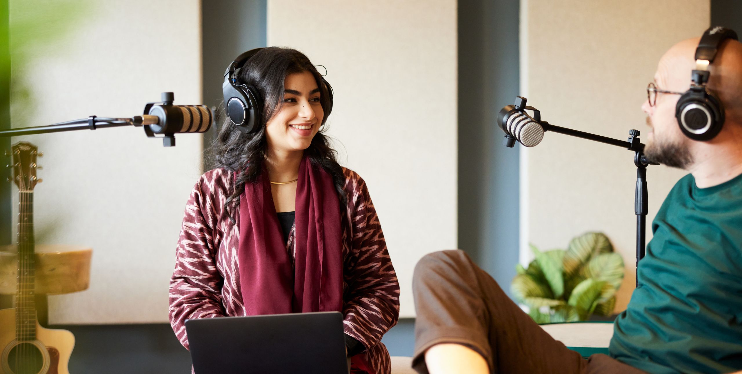 A woman and man with headphones speak into mics for a podcast recording