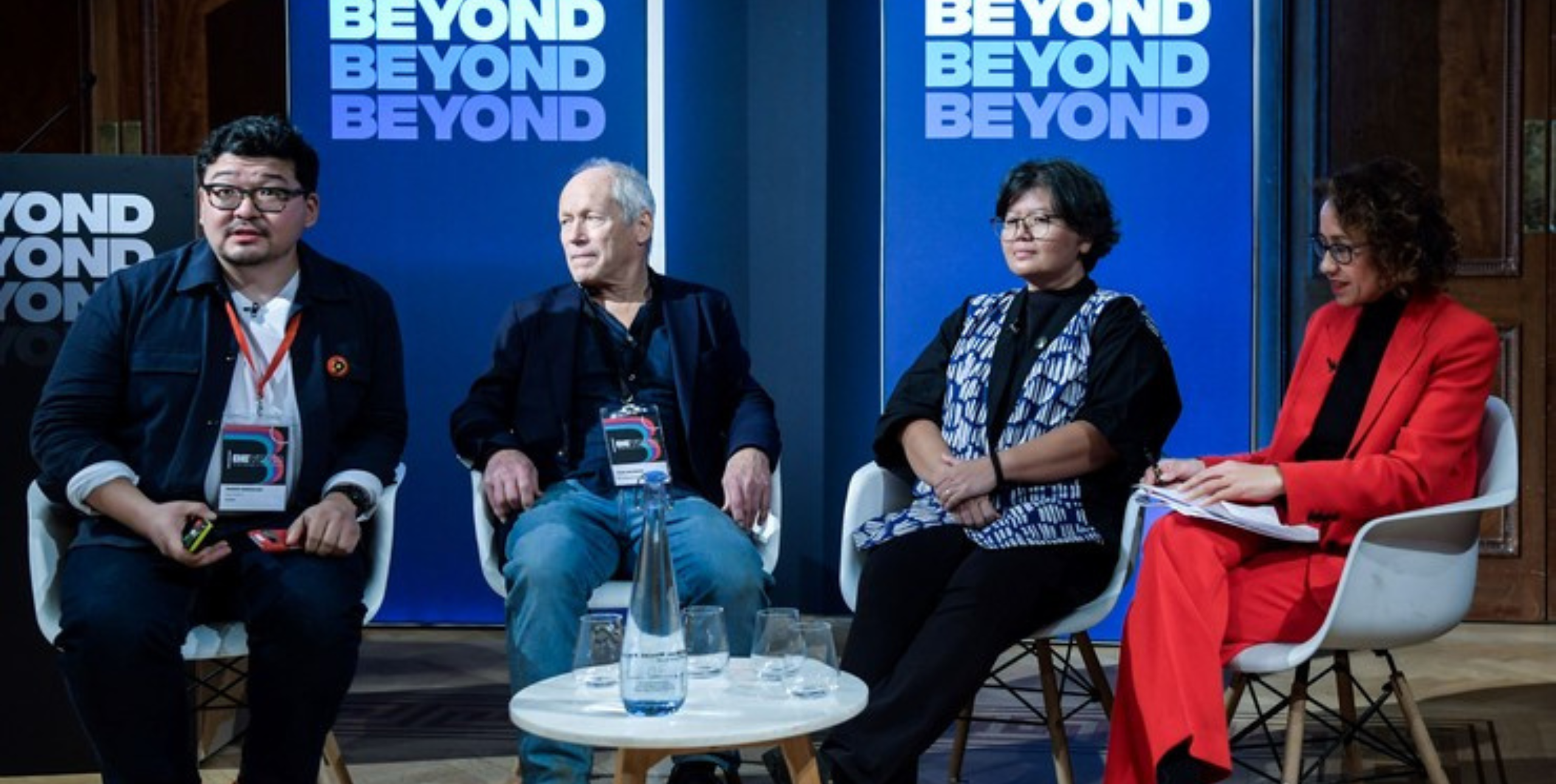 Four people seated on a stage in front of banners reading 'BEYOND' at the BEYOND conference 2023. The people are Daniar Amanaliev, John Newbigin, Tita Larasati and Samira Ahmed.