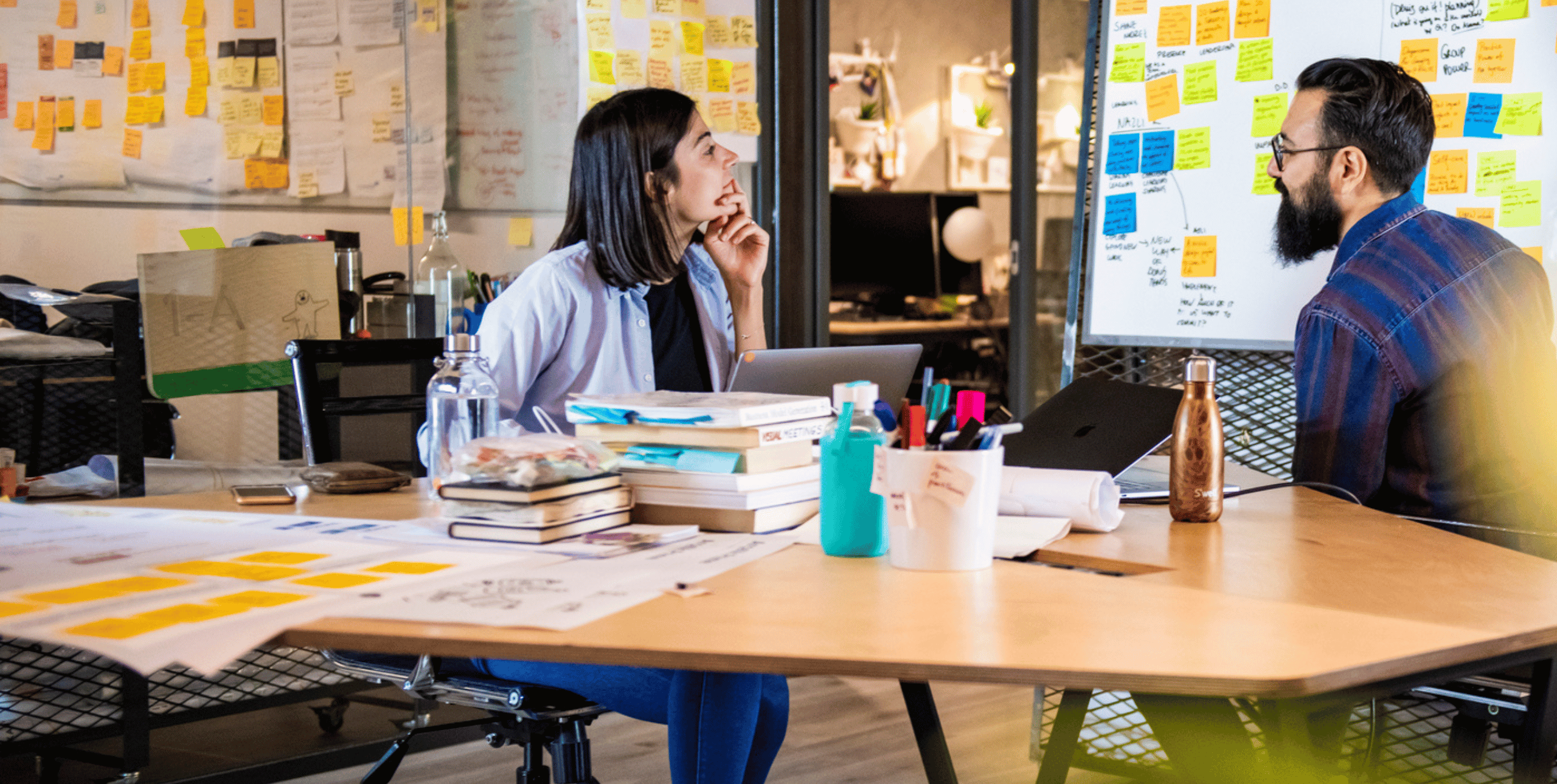 A woman and a man sit at a table in a creative hub. They are chatting and the walls of the room are covered in brightly coloured sticky notes.