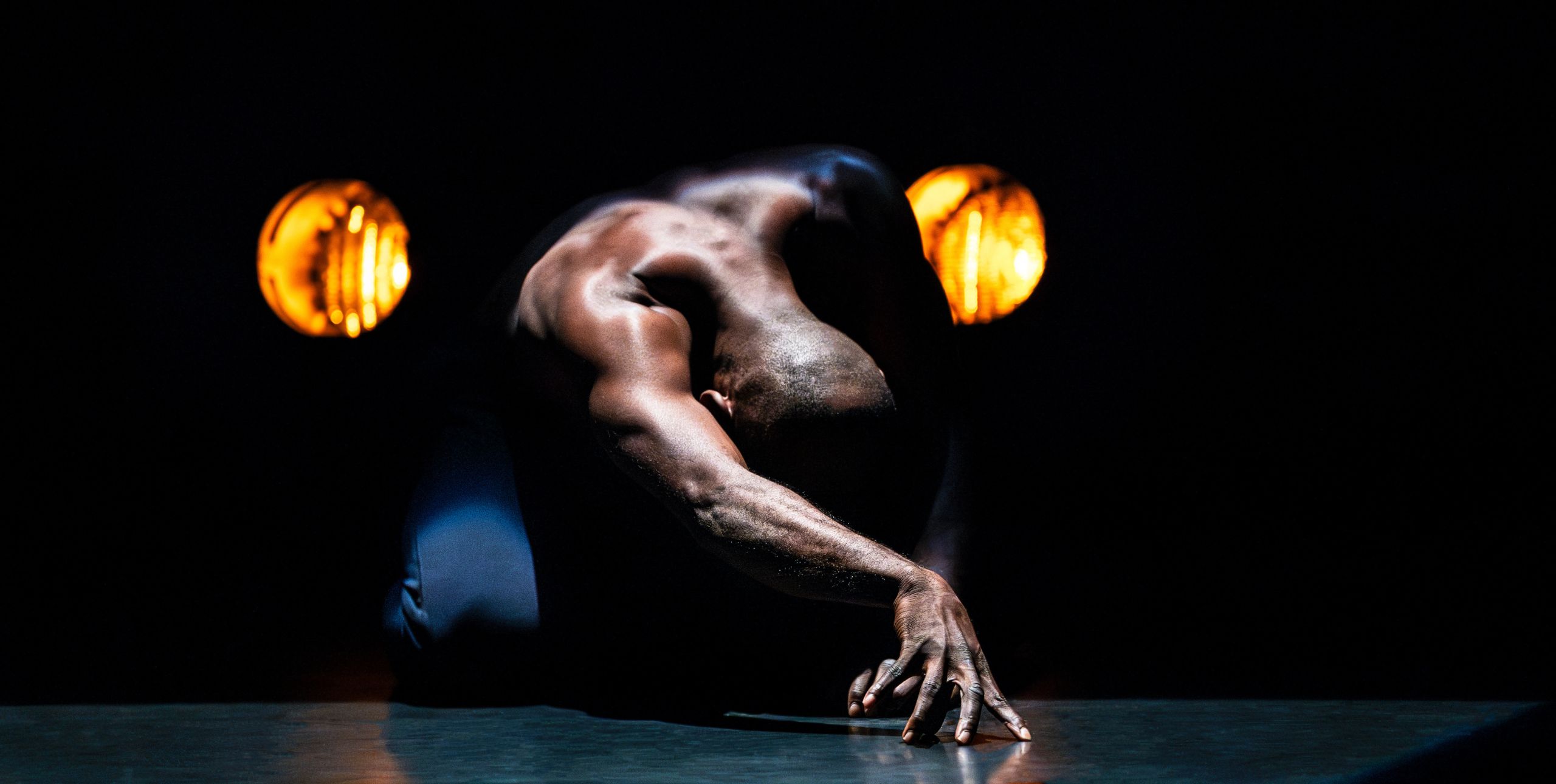 Dancer Enowate kneels on the stage floor in front of car headlights