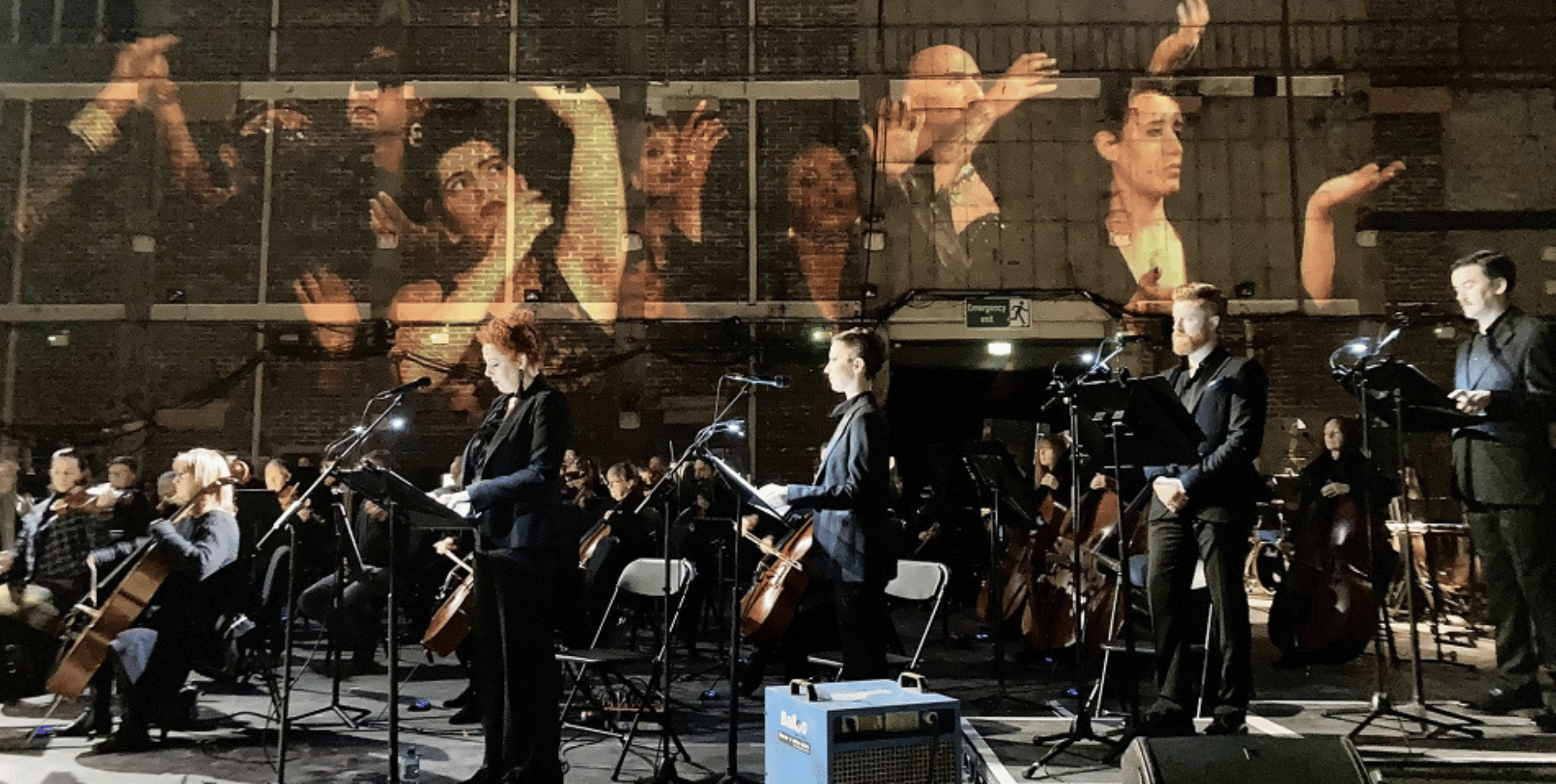 A live performance by the Ulster Orchestra, featuring musicians and vocalists on stage with music stands. The backdrop displays a large projection of expressive individuals with raised hands, creating an engaging visual experience. The setting appears to be an industrial or warehouse-like venue, adding to the unique atmosphere of the event.