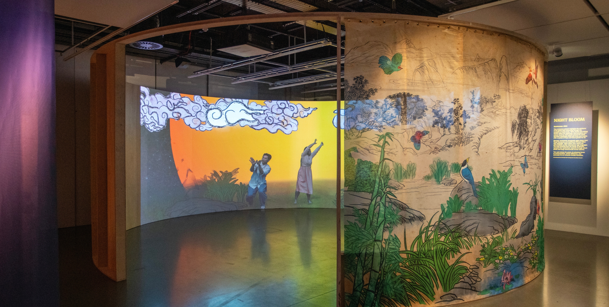 A wooden temporary exhibition at the Science Museum is illustrated with a colourful jungle motif fabric screen. Inside hte installation a vibrant light display shows a landscape and two people waving their arms in the air. 