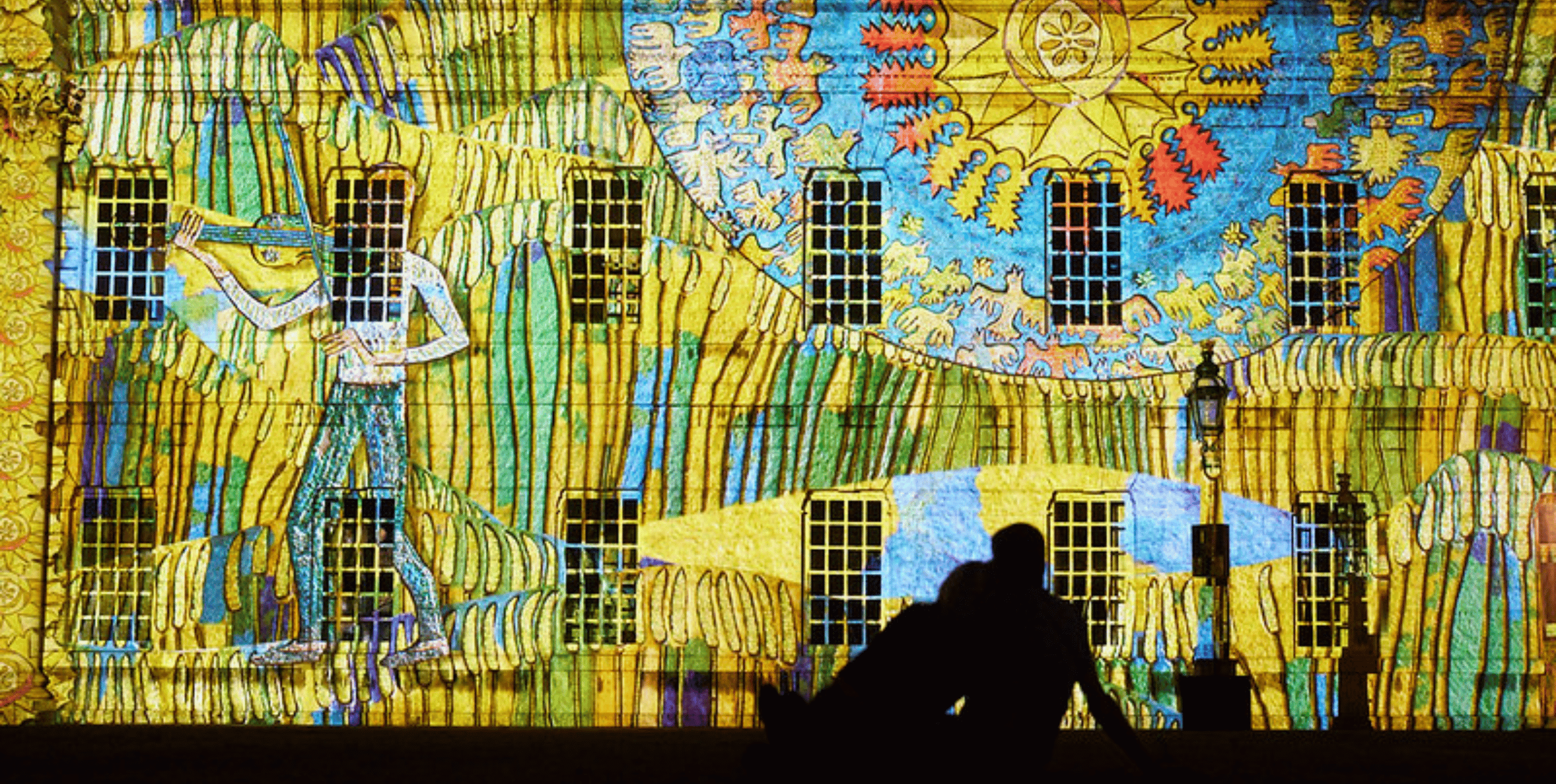 A projection of vibrant blues and yellows in an abstract pattern against the frontage of Buckingham Palace. A seated couple is silhouetted in the foreground, looking on.