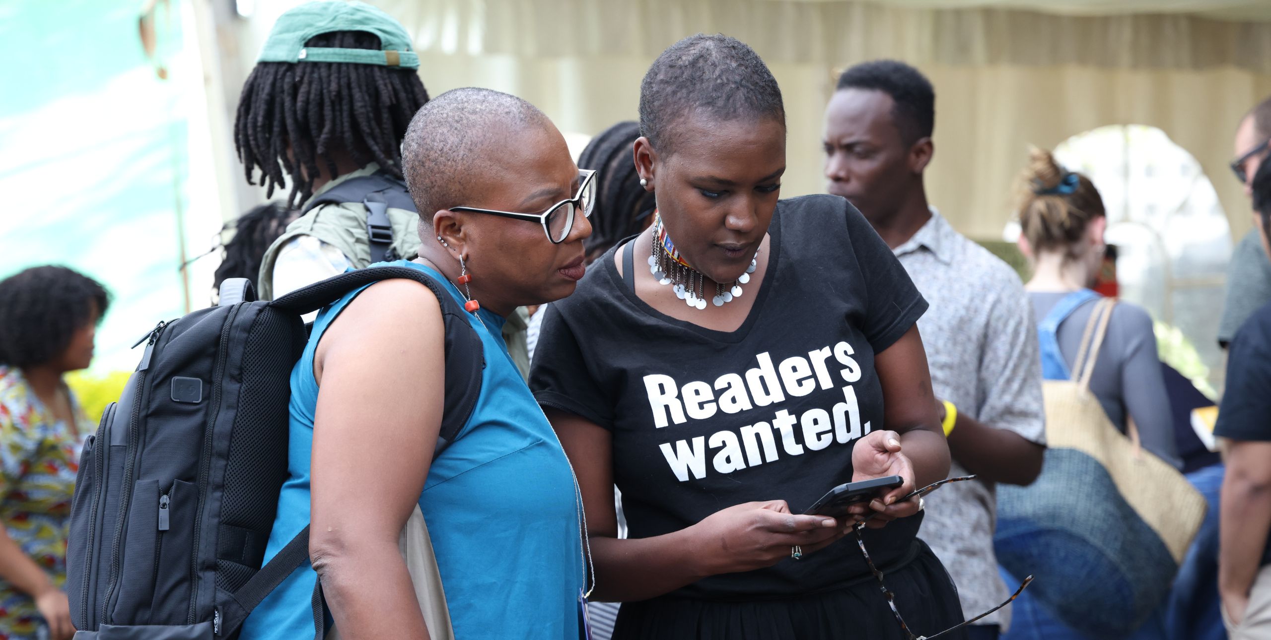 Festival goers, with one wearing a Readers Wanted t-shirt lean over a phone at the NBO Literature Festival 2024