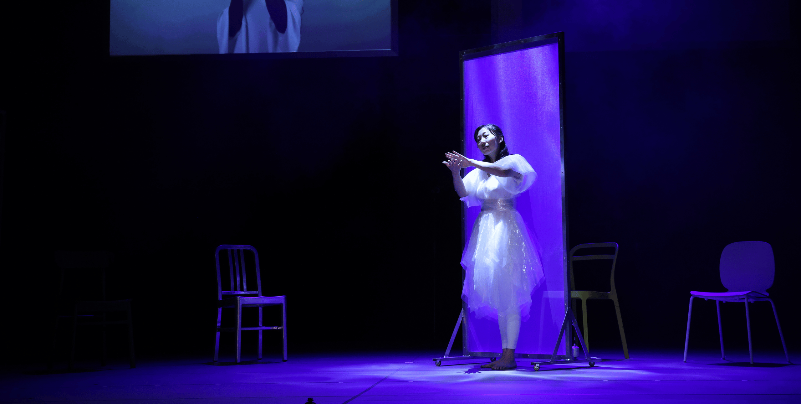 A female performer wearing a white dress stands on a stage dimly lit by blue-purple lighting. She is gesturing towards the audience with her left hand. She stands in front of a tall white screen and there are three chairs arranged around her. At the top left of the frame, the bottom of a screen or projection is just visible, which seems to show the bottom half of a figure in white with long dark hair.