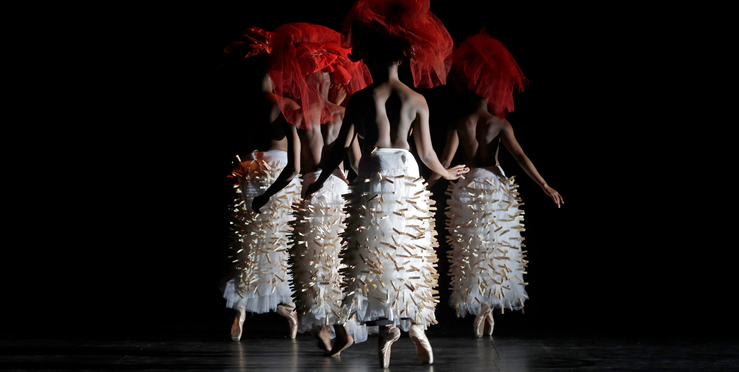 Four performers stand on stage with their backs to the camera. They are topless and wearing ankle-length white tulle skirts with wooden clothes pegs pegged all over the skirts. On their heads they wear red tulle headdresses and they are standing en pointe, wearing pale pink ballet pumps.