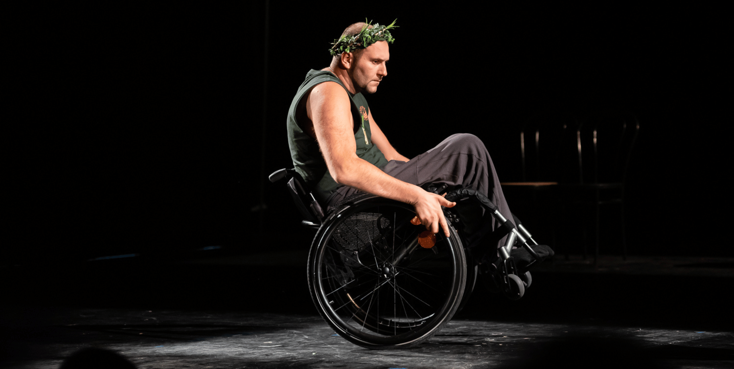 A performer uses a wheelchair and holds in a balance pose with his legs suspended. He is wearing black with a crown made of foliage on his head.