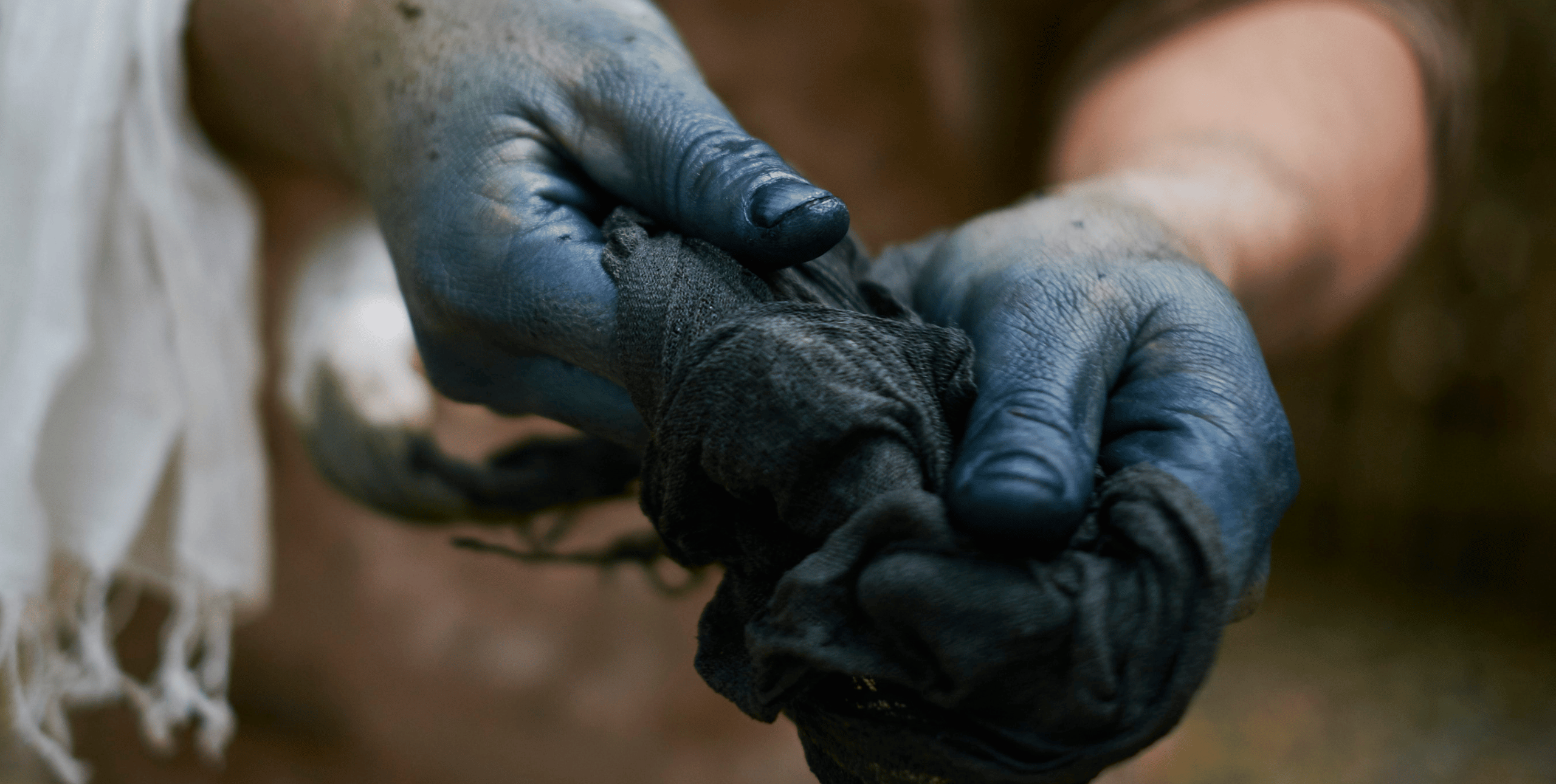 A close up of a pair of hands wring out a dark blue dye-soaked rag. The fingers and thumb are heavily coloured by the dye.