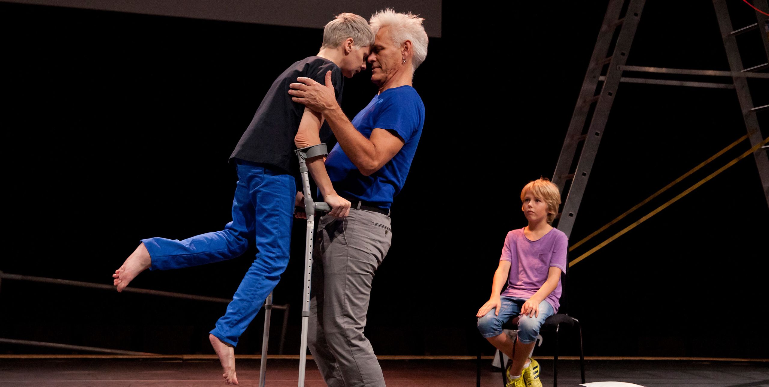 A performer using crutches leans in closely, forehead to forehead, with another adult performer in a tender moment on stage. A child sits nearby on a chair, watching. The scene is part of Claire Cunningham’s The Way You Look (at me) Tonight, performed at Southbank Centre’s Unlimited Festival.