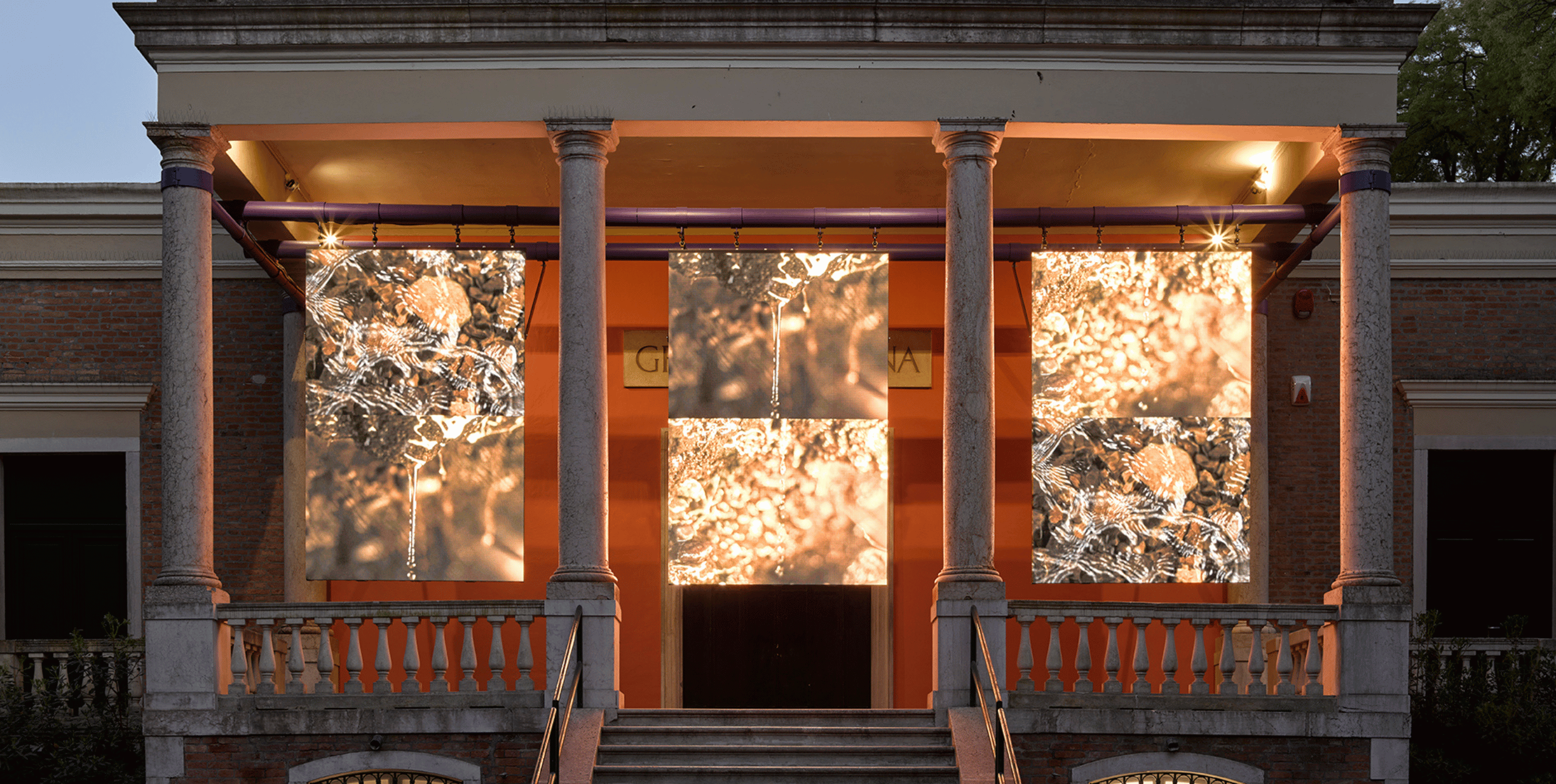 The portico of the British Pavilion building in Venice at dusk. The portico is lit from the rear, with three large screens hanging in the spaces between four pillars. The screens show close ups at different angles of water with sunlight reflecting off its surface. Photo by Jack Hems (c) British Council