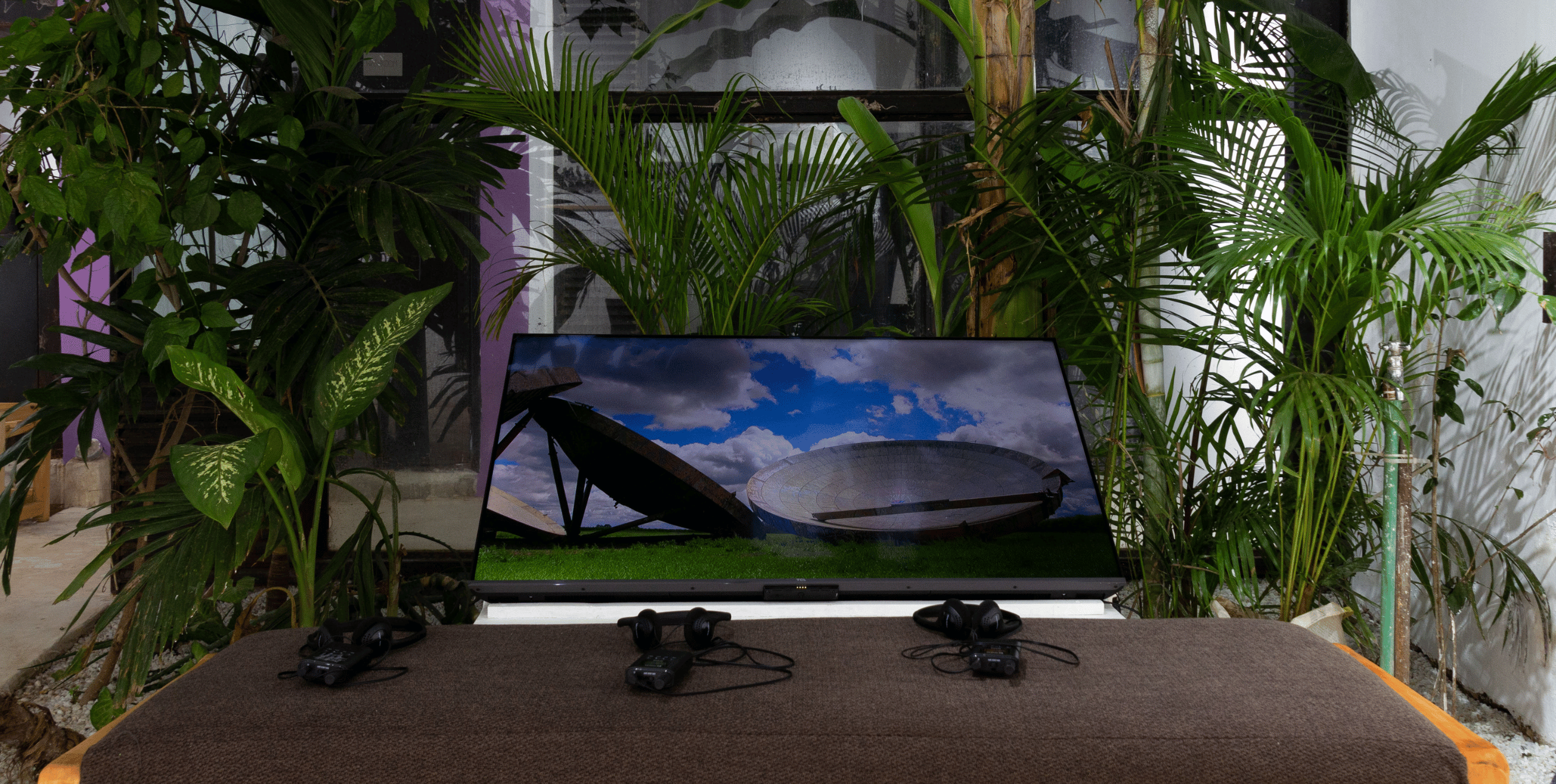 A plasma screen is set on top of a table, angled upwards. The screen shows images of satellite dishes against a bright blue sky with white clouds. In front of the screen there are headphone sets, and around the display are tropical leaved plants.