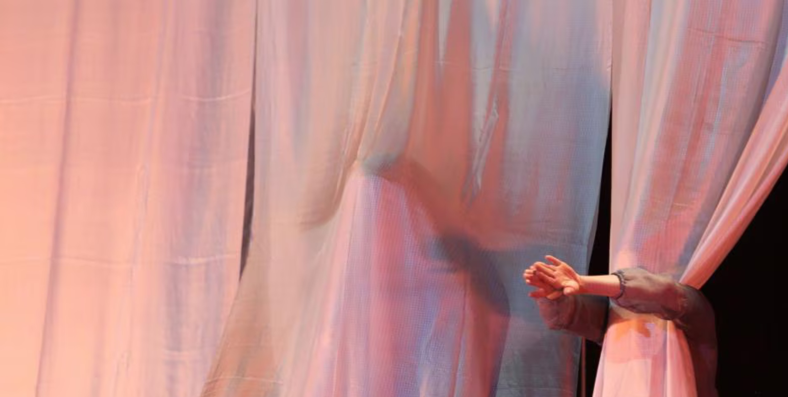 A person stands behind a translucent pink curtain infront of an unlit stage, their head is hidden behind the curtains but their hands clasp together around the front