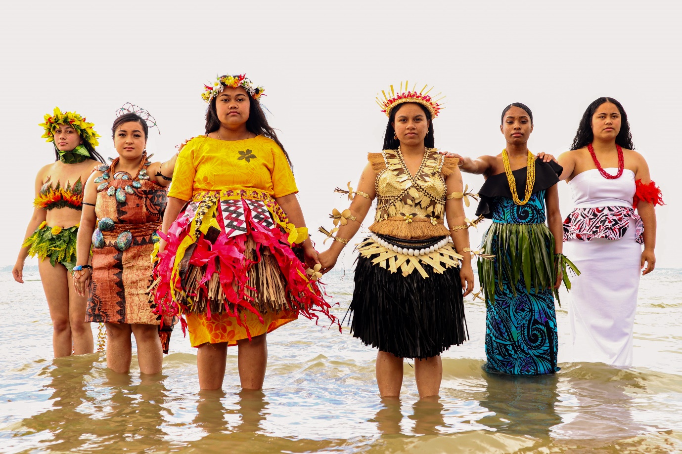Six people in ceremonial dress stand in shallow seawater, wearing woven garments, shells, floral adornments and headdresses, with the sea and sky in the background.