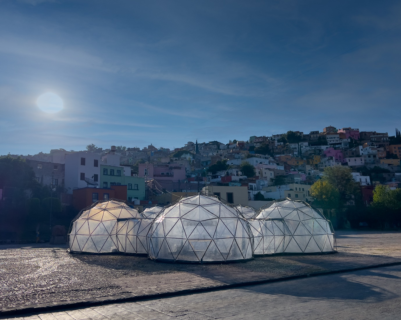Transparent geodesic domes stand in a public square beneath a hillside cityscape under a bright blue sky.