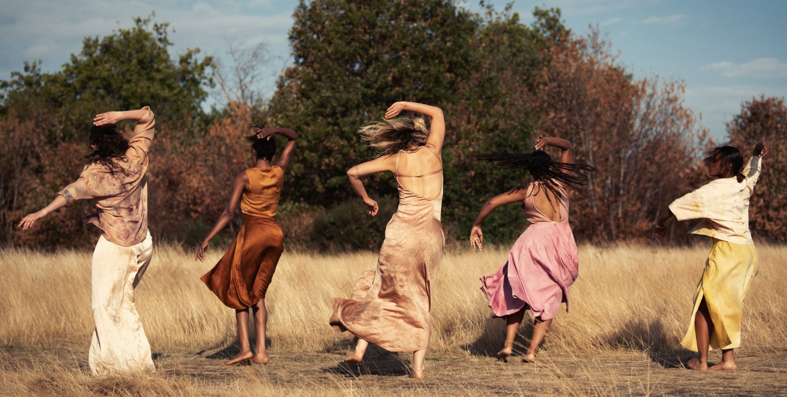 Five women in flowing dresses dance outside in nature