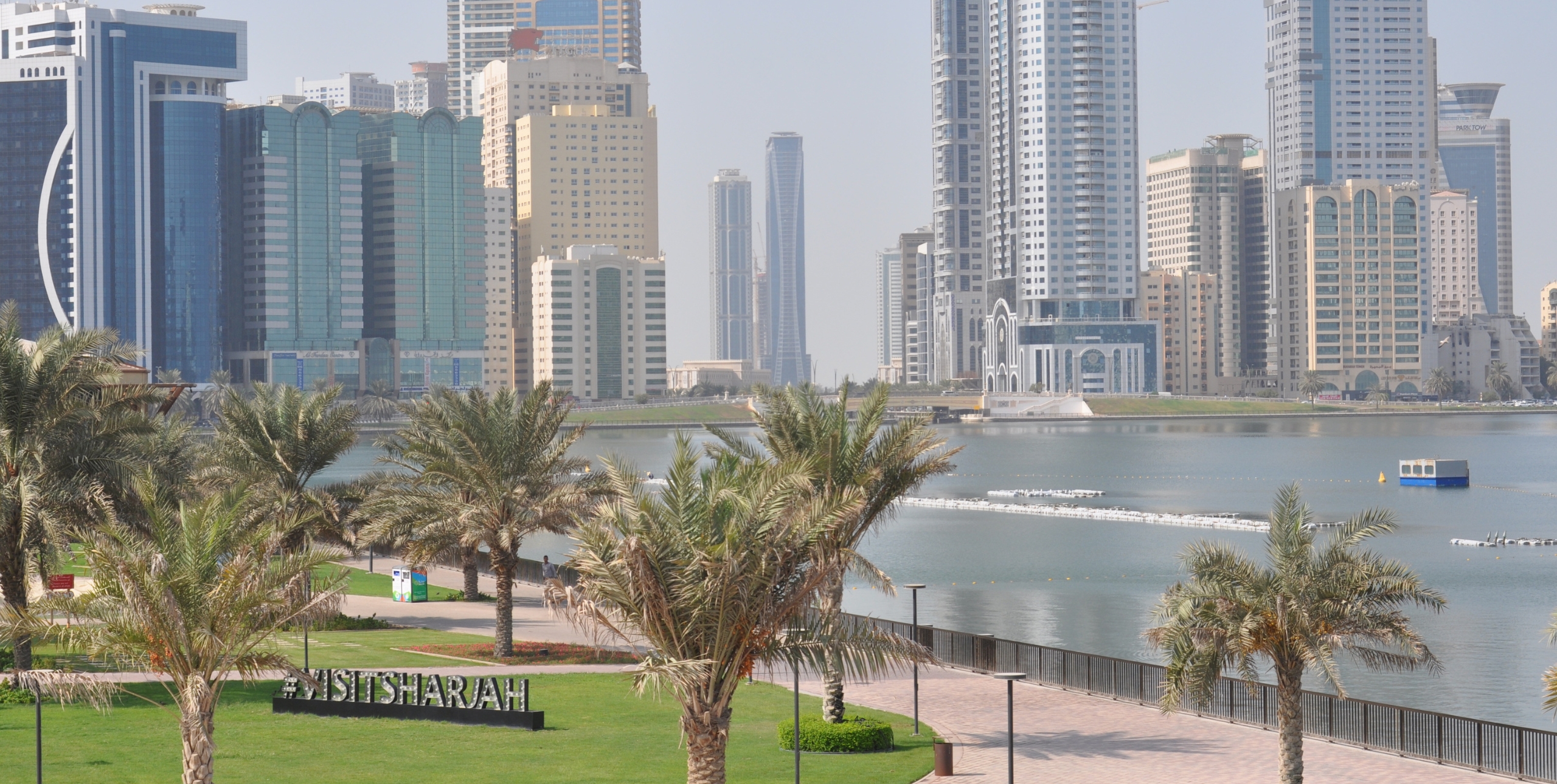 A waterfront view of Sharjah’s modern skyline, with palm-lined promenades in the foreground, sparkling calm water and high-rise buildings behind, and a “#VISITSHARJAH” sign nestled among the palms.