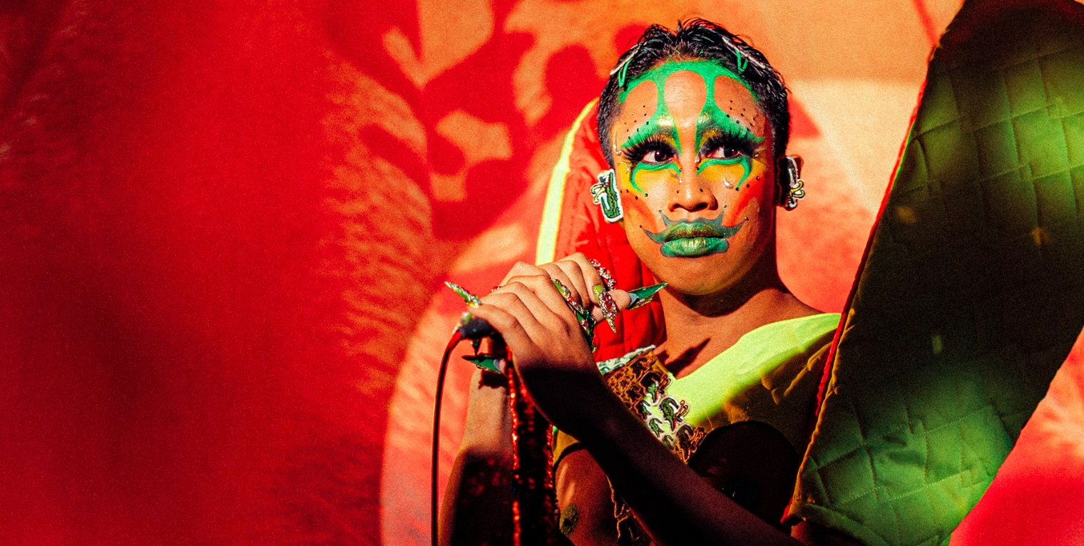 A performer in Filipino traditional dress and bright green face paints stands on stage holding a microphone on a stand, and looks out toward the right of the image