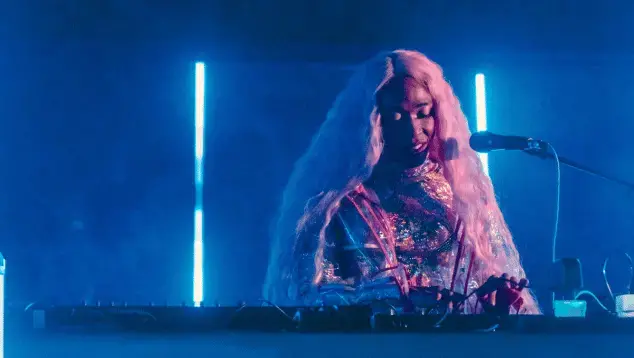 A Black woman, wearing a sequined outfit and with long pink hair, stands in front of a microphone and operates a sound desk. Behind her is a haze of dry ice and bright blue neon lights.