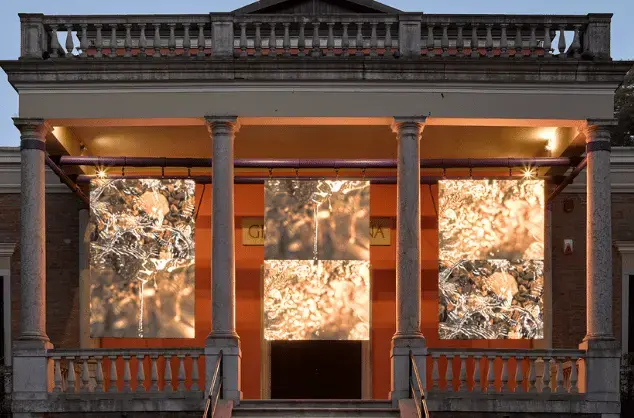 The portico of the British Pavilion building in Venice at dusk. The portico is lit from the rear, with three large screens hanging in the spaces between four pillars. The screens show close ups at different angles of water with sunlight reflecting off its surface. Photo by Jack Hems (c) British Council