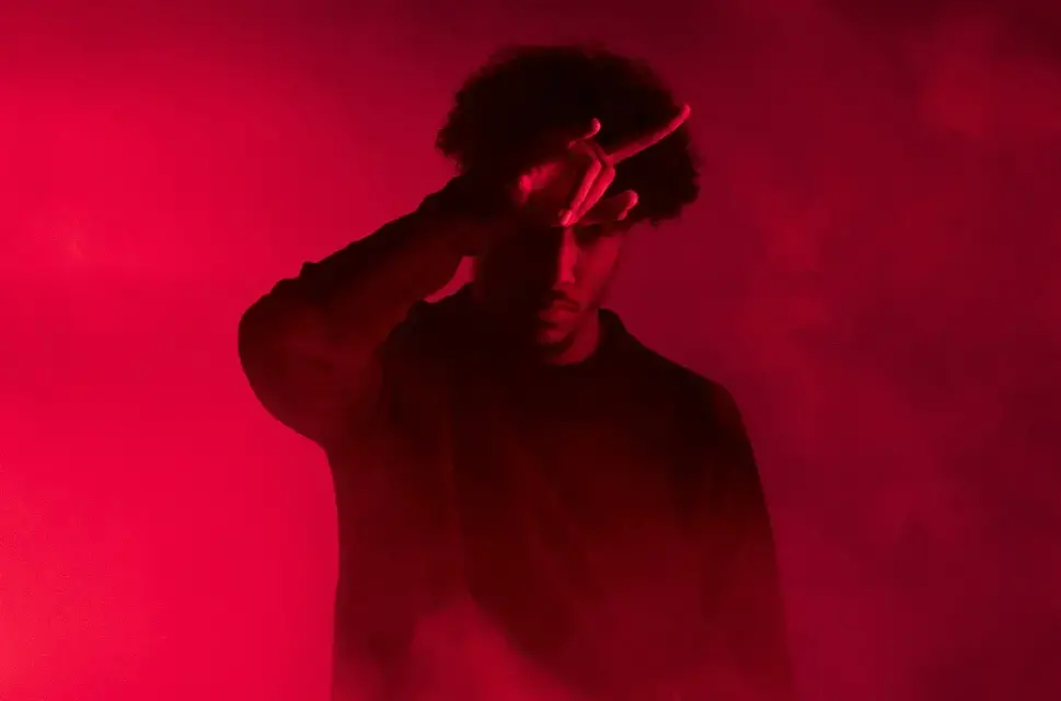 Man with hand against his head pointing two fingers out, against a dark red, smoky background.