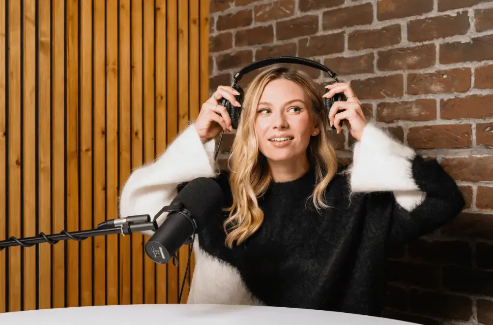 Sian Eleri, Selector Radio presenter. A woman with blonde hair sits in a radio studio, smiling, behind a large microphone, holding headphones.