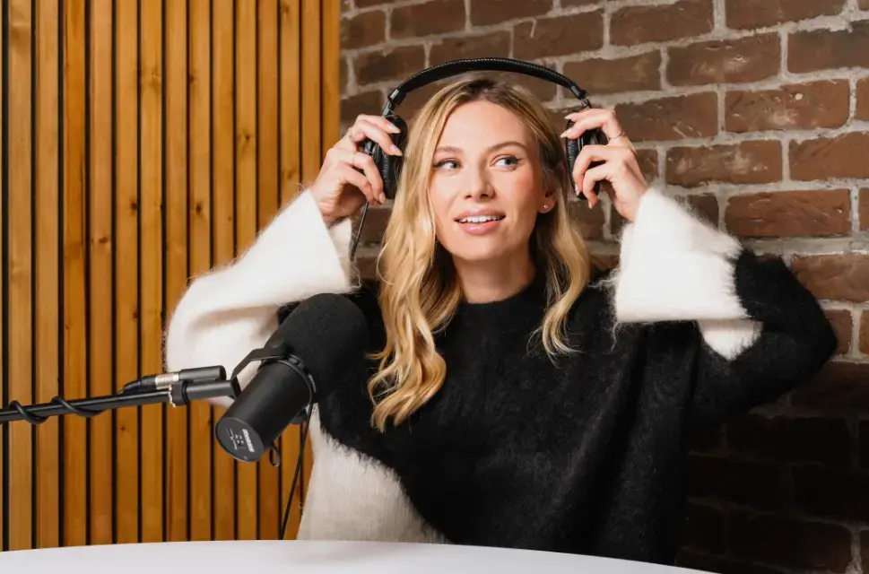 Lady in radio studio, smiling, behind a large microphone, holding headphones.