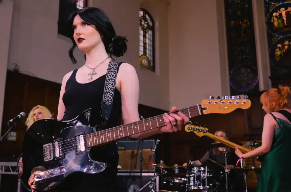 Band on stage in church, lady at the front holding her electric guitar, looking forwards.