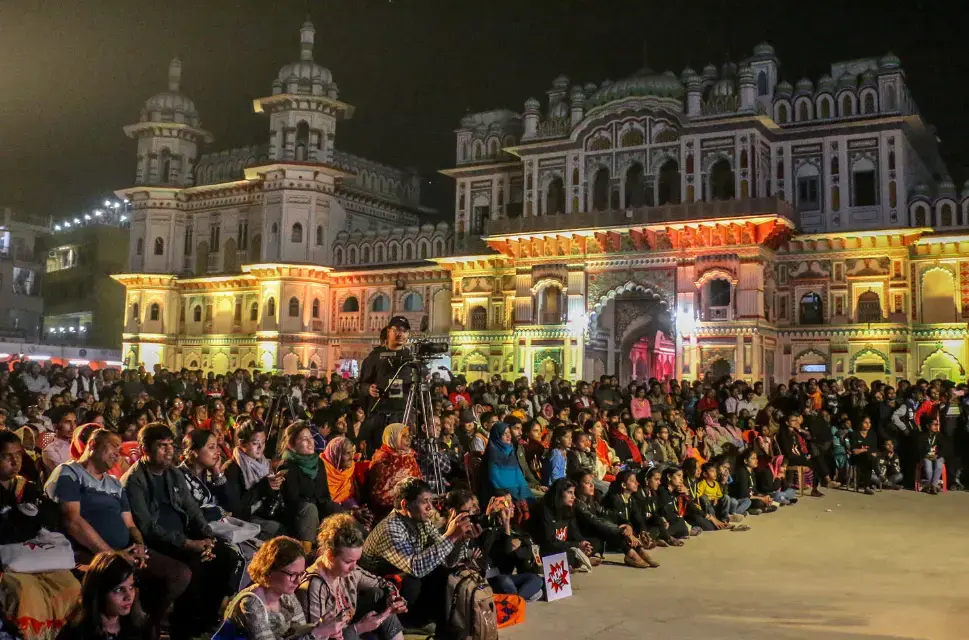 A crowd of people seated on the floor watch a nighttime event at WOW Nepal in 2019. The crowd is seated in front of a grand building lit up by multicoloured lights. The event they are watching is taking place outside of the frame of the image, to the right.