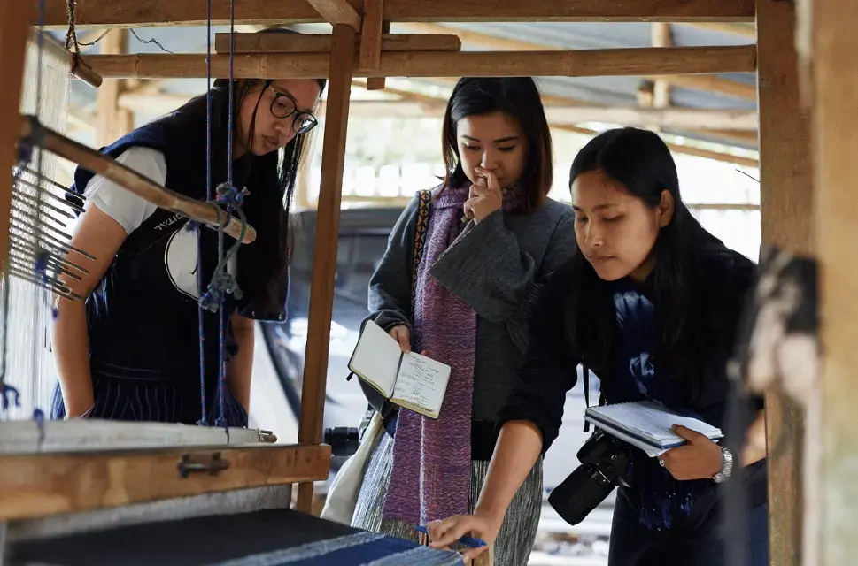 Three young women examine a manual fabric weaving loom, holding notepads. Crafting Futures, Thailand, photography by Simon Mills