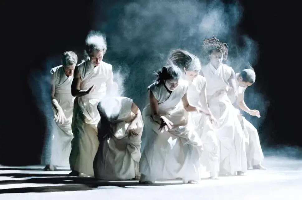 Seven performers stand on stage surrounded by white smoke or a cloud of powder. They are dressed in white and are gesturing with their hands, with some standing and some crouching slightly.Photo by Richard Haughton