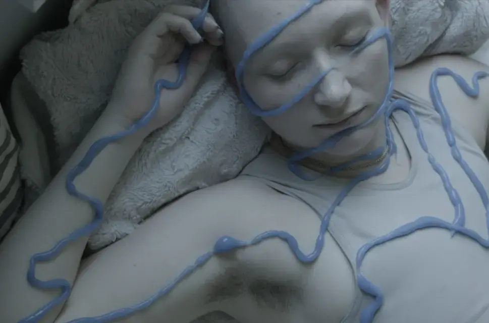 A woman lies on a soft bed in a flesh coloured top. Plastic looking blue liquid zig zags across her face, arms and torsoe.