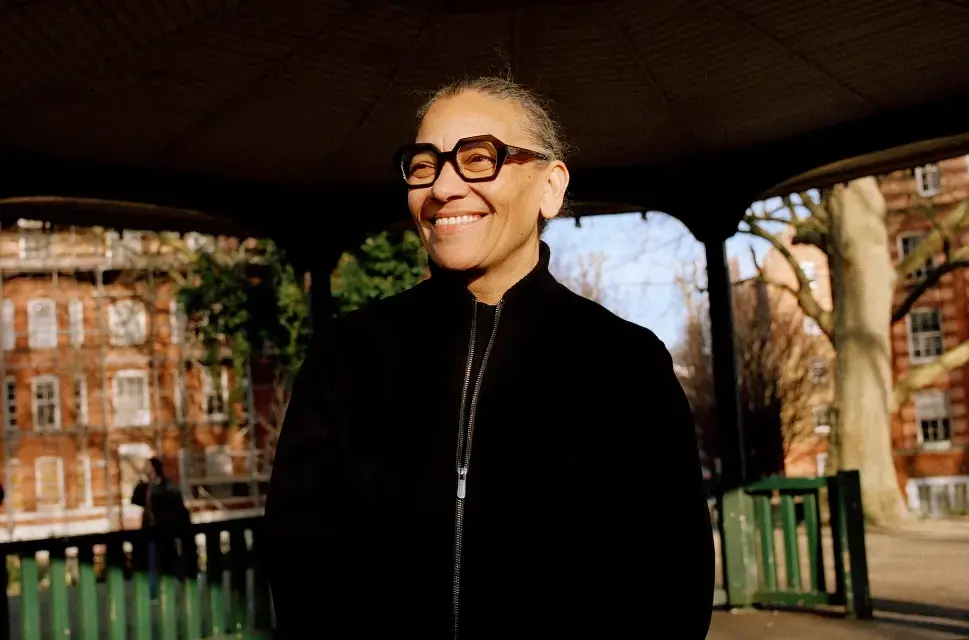 Lady outside, smiling, wearing a black zip-up jumper, black glasses, stood under the cover of a bandstand with large trees and buildings behind her.