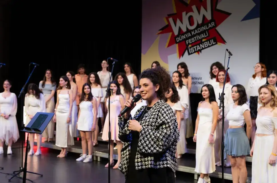 A woman stands on stage infront of a choir of women dresesd in white as part of WOW Istanbul 2024.