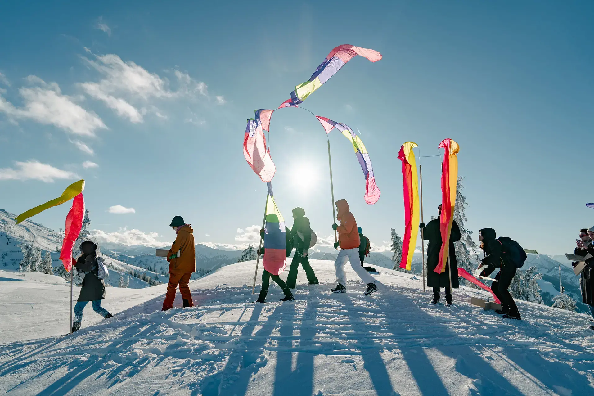 A line of people walk along a snow filled mountain landscape waving long colourful flags as part of the minus 20 Degrees festival, Austria