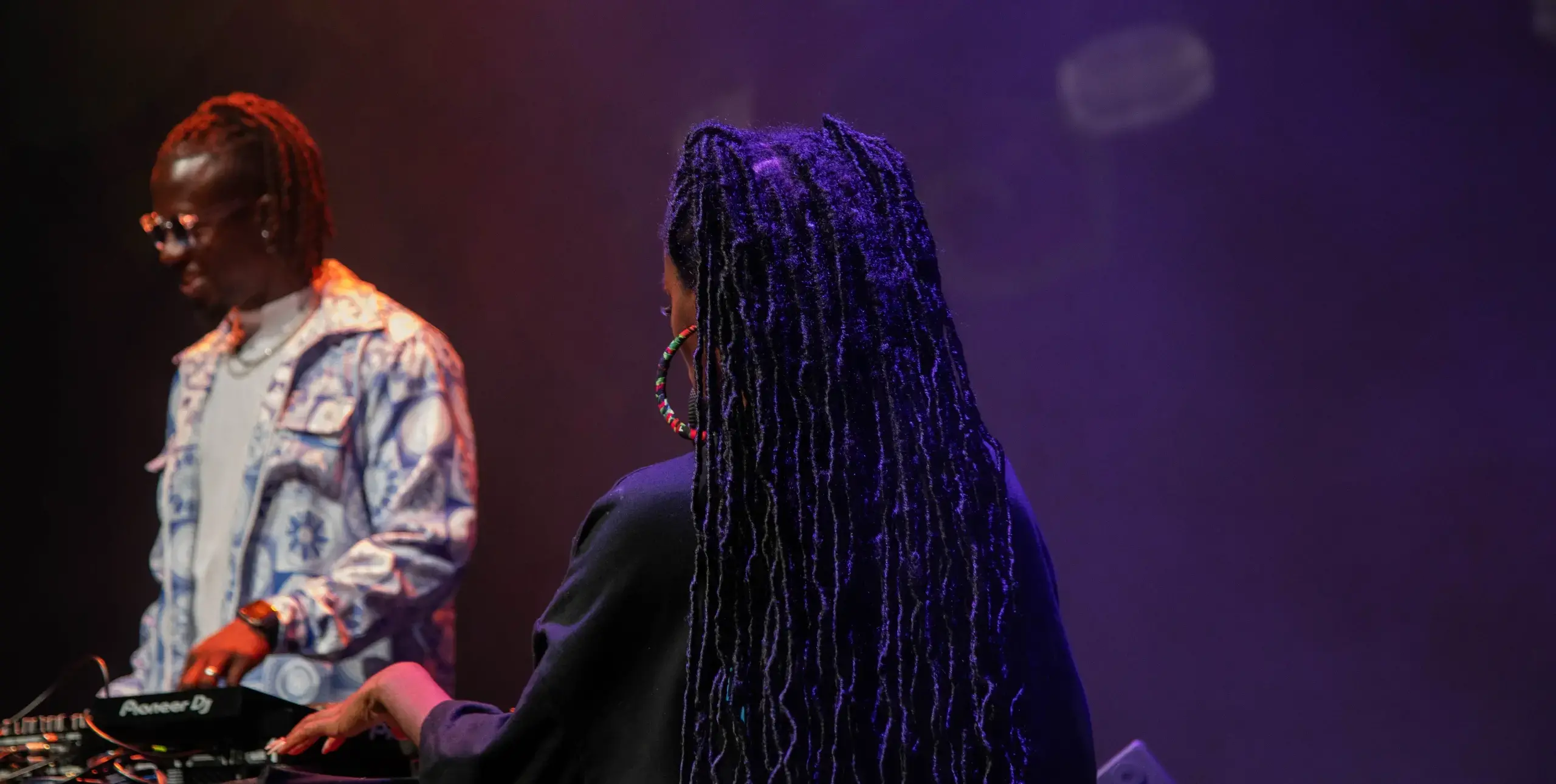 A man in the background DJs on a dark stage at the Afrodiaspora festival in Columbia, centered in the photo is the back of a woman with long braided hair holding a microphone on stage