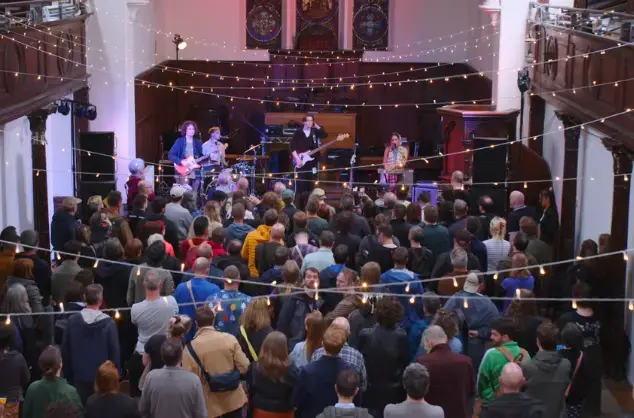 Standing crowd in coverted church facing four-piece band on stage. There are small lights hanging from lines above them.
