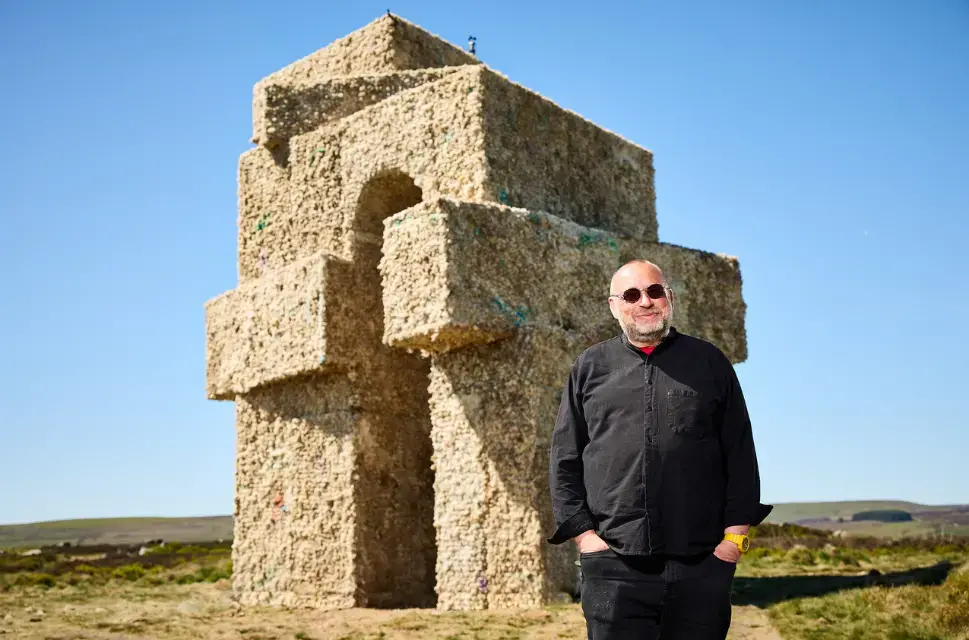 Man smiling, wearing all black including his sunglasses, in front of a large tower in the countryside.