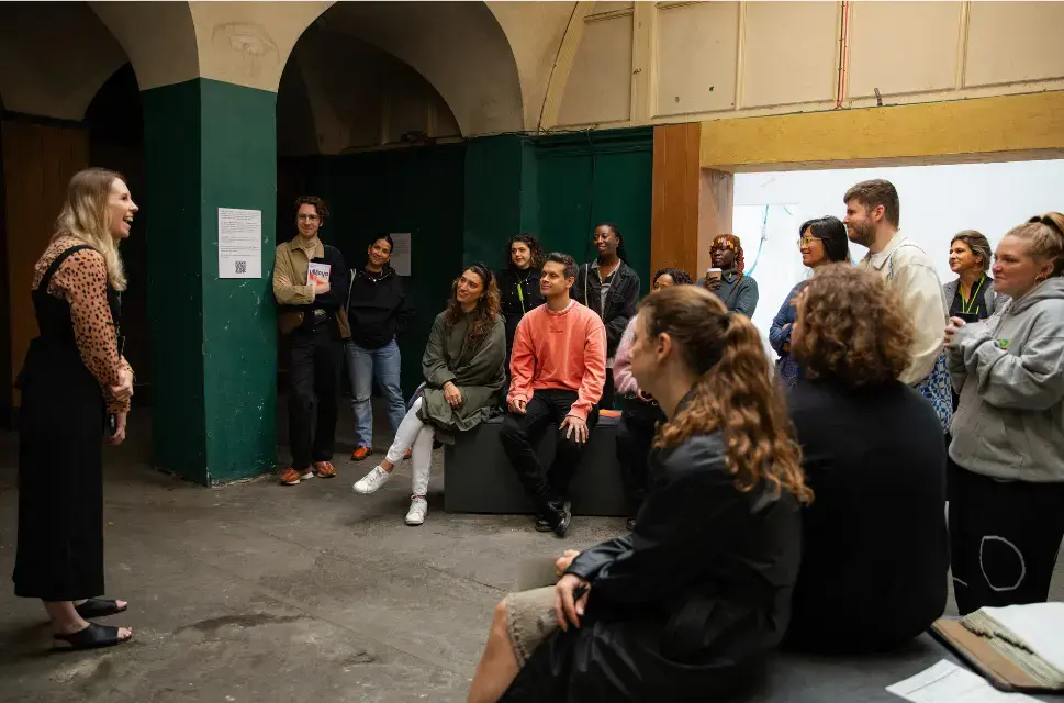 Group of people in a side room at an exhibition with a lady in the middle of the group, hosting and smiling.