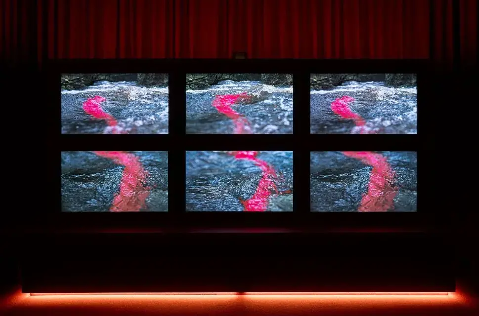A still from John Akomfrah's Listening All Night To The Rain featuring 6 video screens with red swirl on a textured grey background