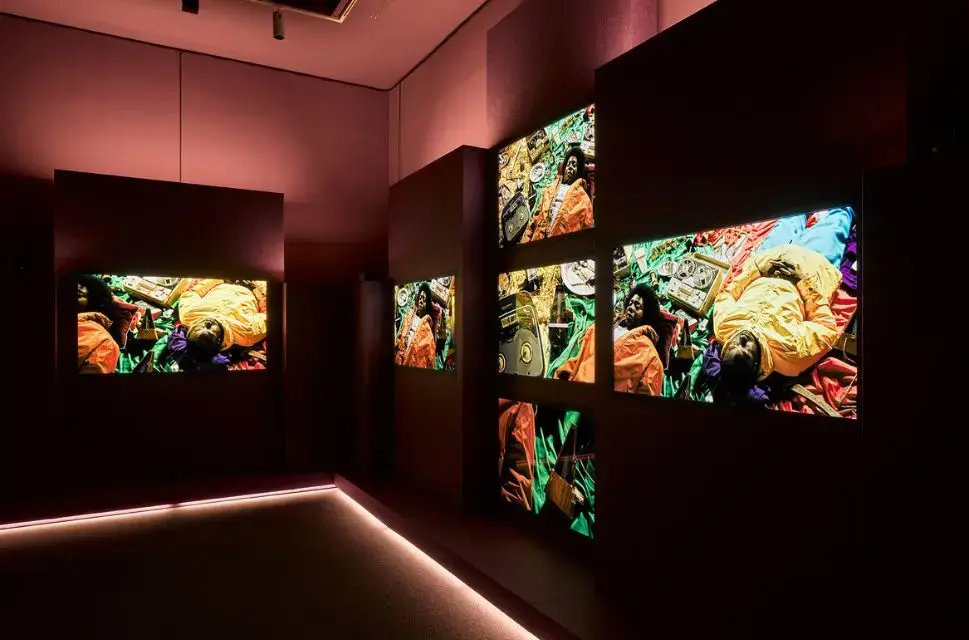 A still from John Akomfrah's Listening All Night To The Rain featuring 6 video screens with a scene of people lying on colourful rugs