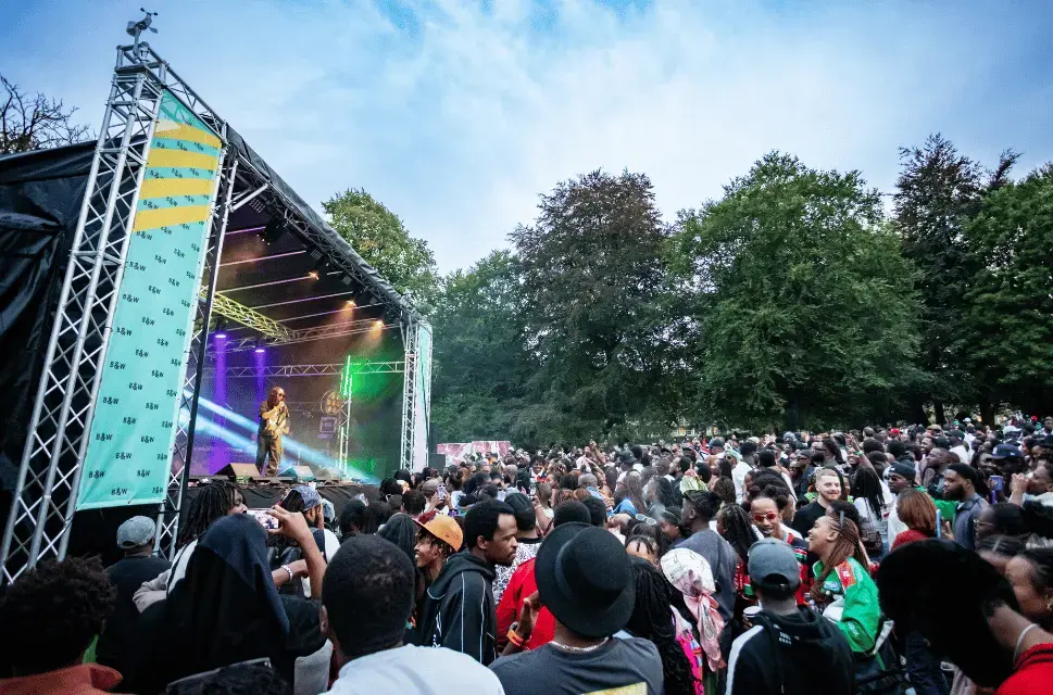 A crowd stands in front of an outdoor concert stage, with bright lights coming from the stage and blue sky above. There is a lone performer in the centre of the stage. Blankets & Wine, part of Bradford City of Culture 2025. Photo by Andrew Benge.