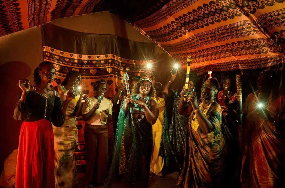 A group of women dance together in a tent-like structure. They are wearing bright colours and hold instruments and mobile phones with torches activated over their heads. Storytelling as Safeguarding: Protecting South Sudanese Women's Cultural Heritage in Refugee Settings in Uganda and Kenya. Photo by Likikiri Collective