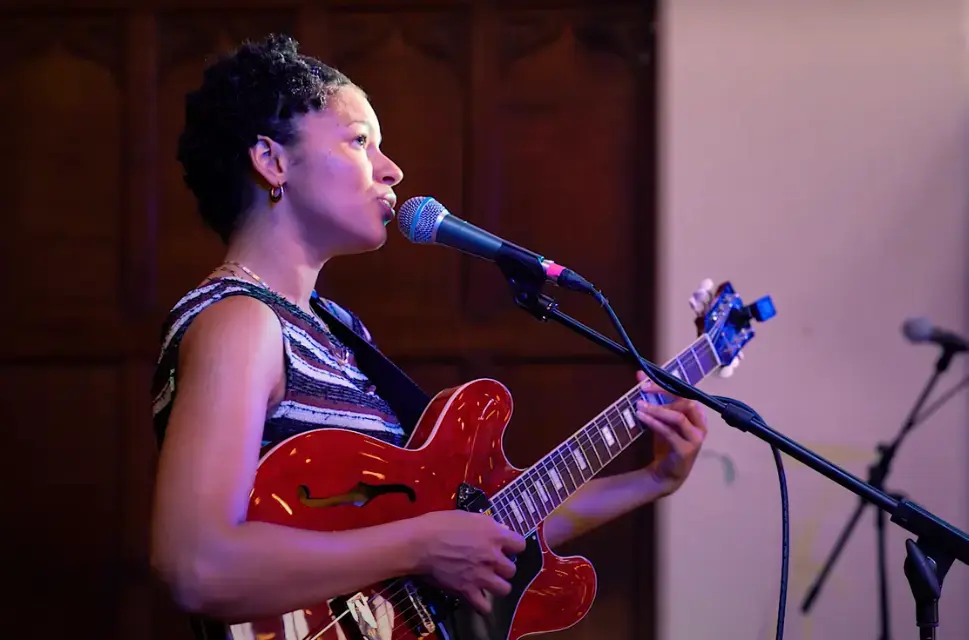 Side on of lady holding guitar, singing into microphone on a stand.