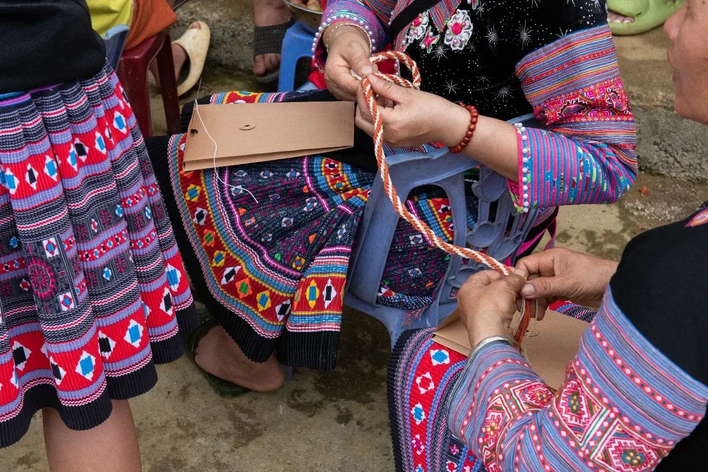 Close-up of people in brightly patterned traditional clothing weaving or braiding cord while seated outdoors.