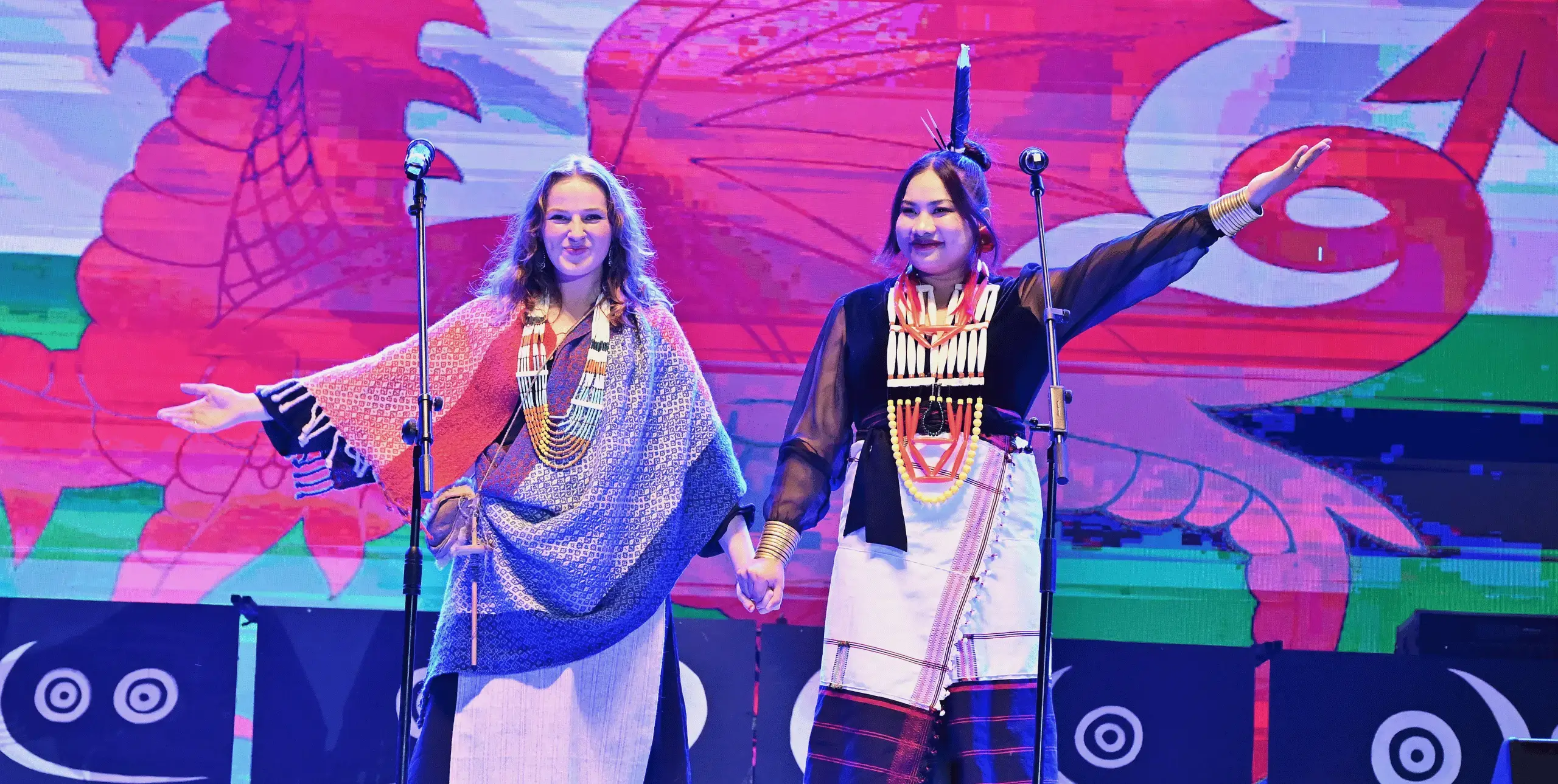 Two women stand on stage looking out to an audience out of shot. They are holding hands and waving to the audience, smiling. They stand against a large Welsh flag and are wearing traditional Indian clothing.