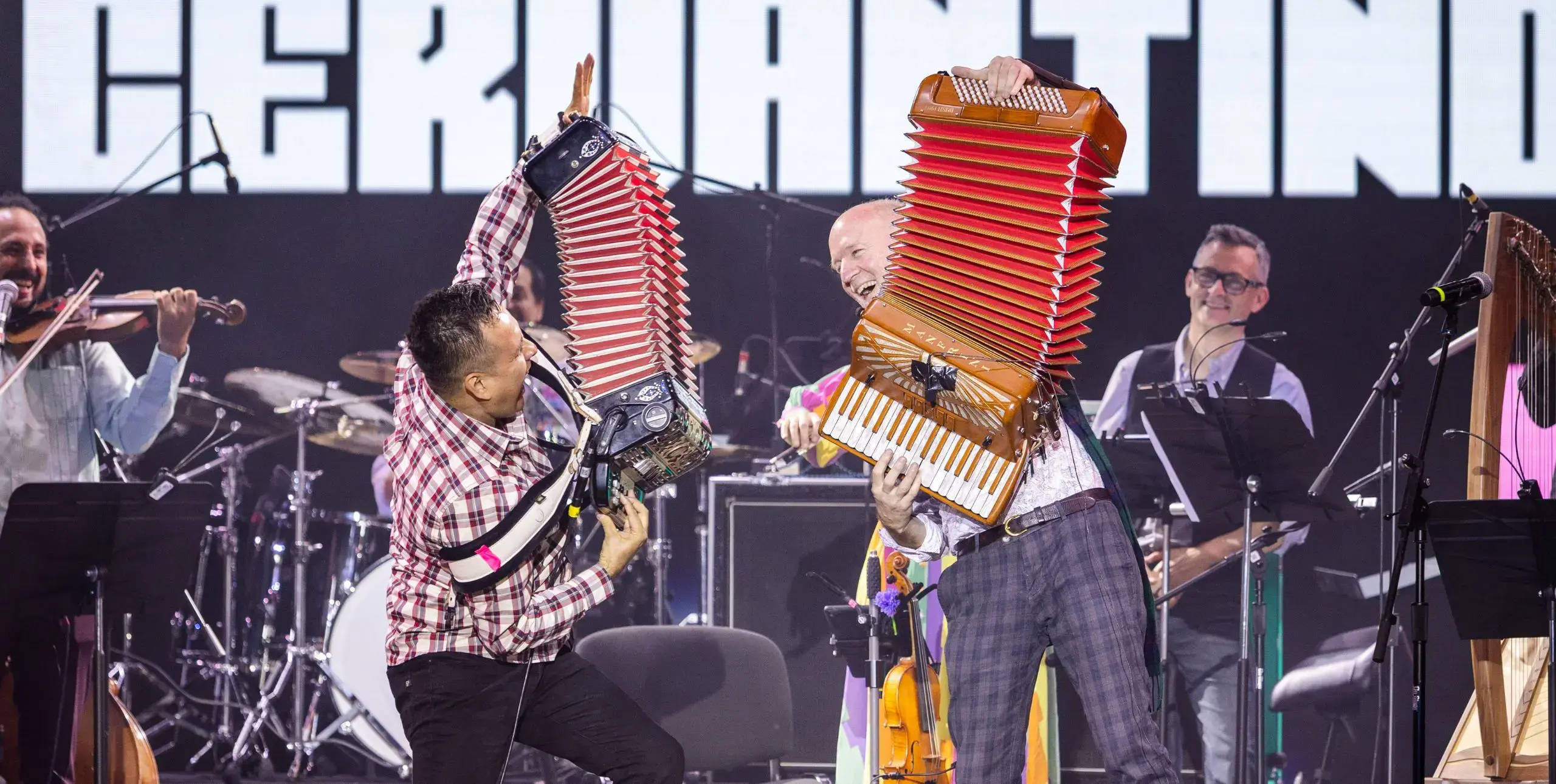 Two musicians, one Scottish and one Mexican, play the accordian energetically on a stage with the rest of the band looking on