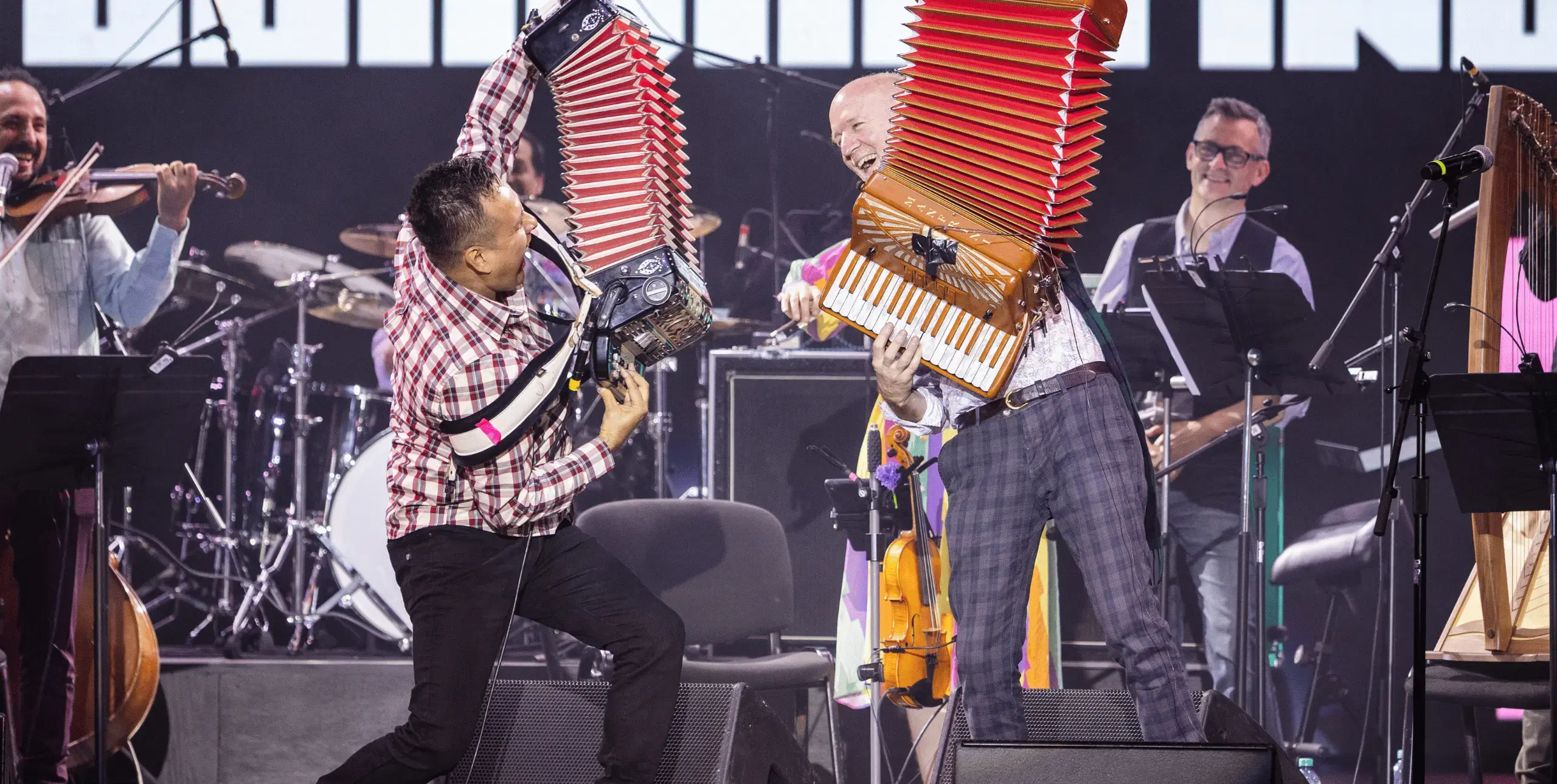 Two musicians playing accordions on stage, facing each other in dynamic poses, with joyful expressions on their faces. Other musicians are seen in the background, and a violinist is seen to the left of the frame.