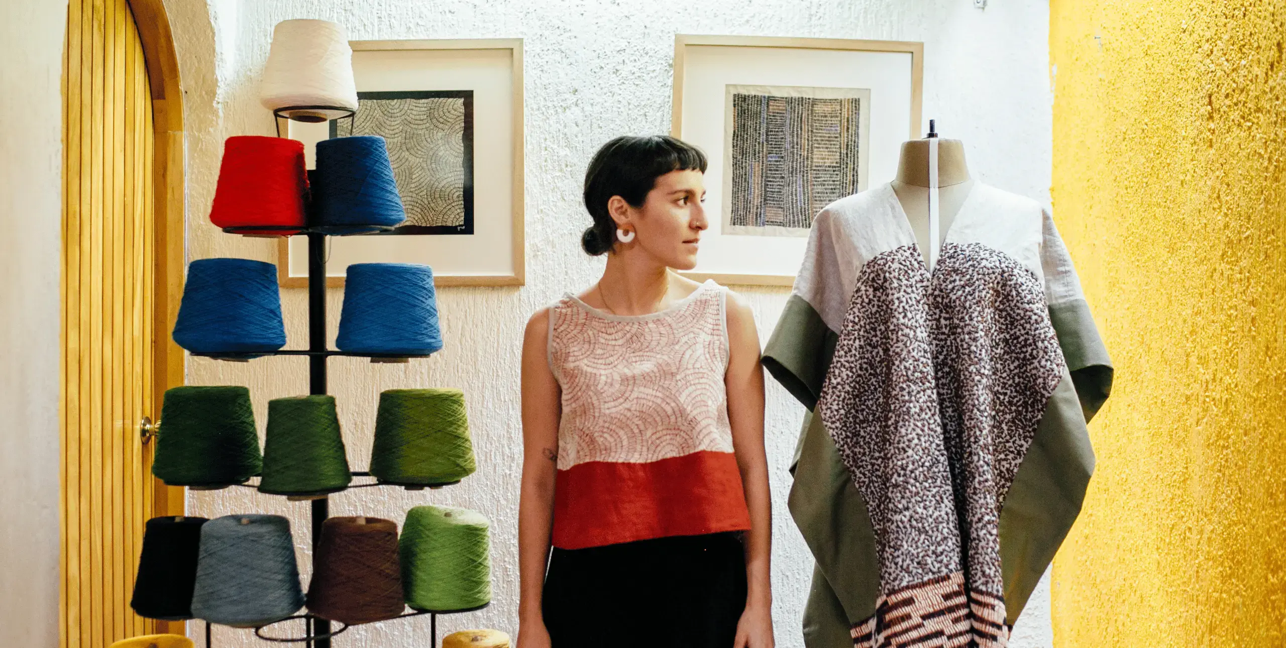 A woman stands between a manequin and a stand holding spools of coloured yarn. She looks to her left, beyond the manequin. She's wearing a gold earrings and a black, white and red striped top, her short dark hair is tied back. There are two framed pictures out of focus hanging on the wall behind her.