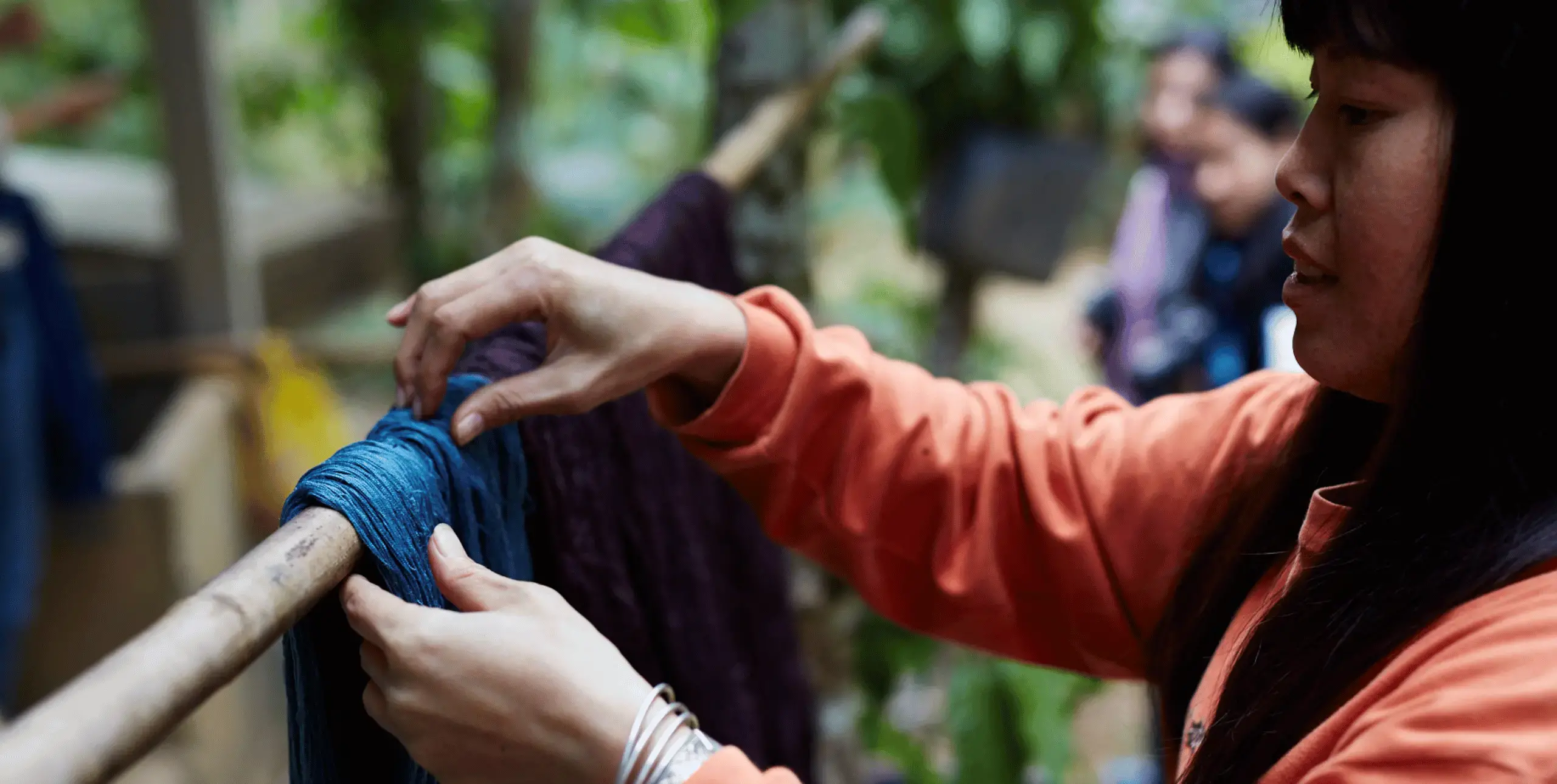 A woman hangs freshly dyed fabric over a pole suspended between trees in a woodland.